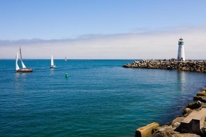 Lighthouse on rocky coast with sailboats on ocean under blue sky.